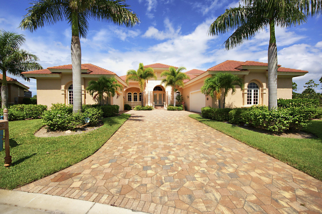 Majestic beige house with a red-tiled roof, bordered by lush greenery and tall palm trees. A paved driveway leads to an elegant entrance. Bright, sunny day.