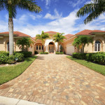 Majestic beige house with a red-tiled roof, bordered by lush greenery and tall palm trees. A paved driveway leads to an elegant entrance. Bright, sunny day.