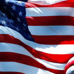 The image shows a close-up of the U.S. flag, with vibrant red, white, and blue colors. The fabric waves dynamically, conveying a sense of pride and motion.